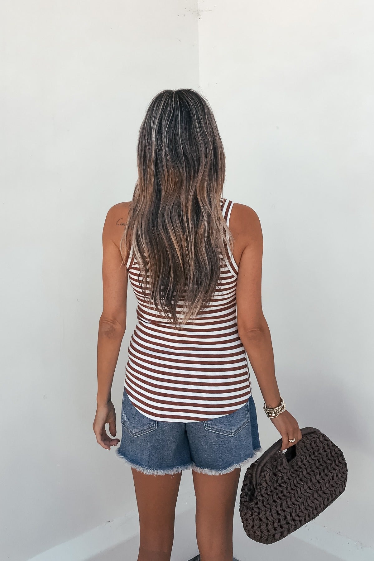 A woman with long wavy hair in a Lorna Stripe Racerback Tank Top - Brick and denim shorts stands holding a textured clutch by a white wall.