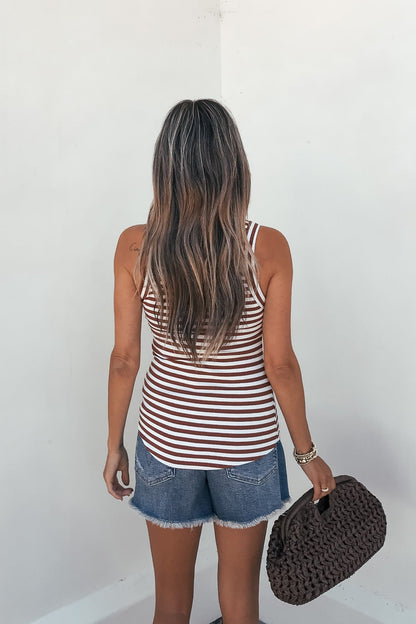 A woman with long wavy hair in a Lorna Stripe Racerback Tank Top - Brick and denim shorts stands holding a textured clutch by a white wall.