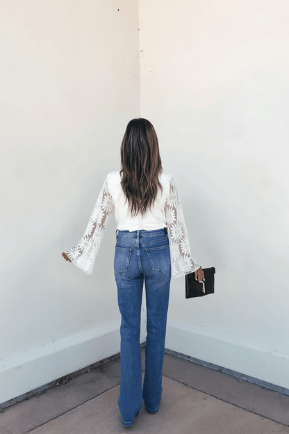 Woman facing a white wall, wearing flared jeans, the Louisa White Floral Lace Bodysuit, and holding a black clutch.