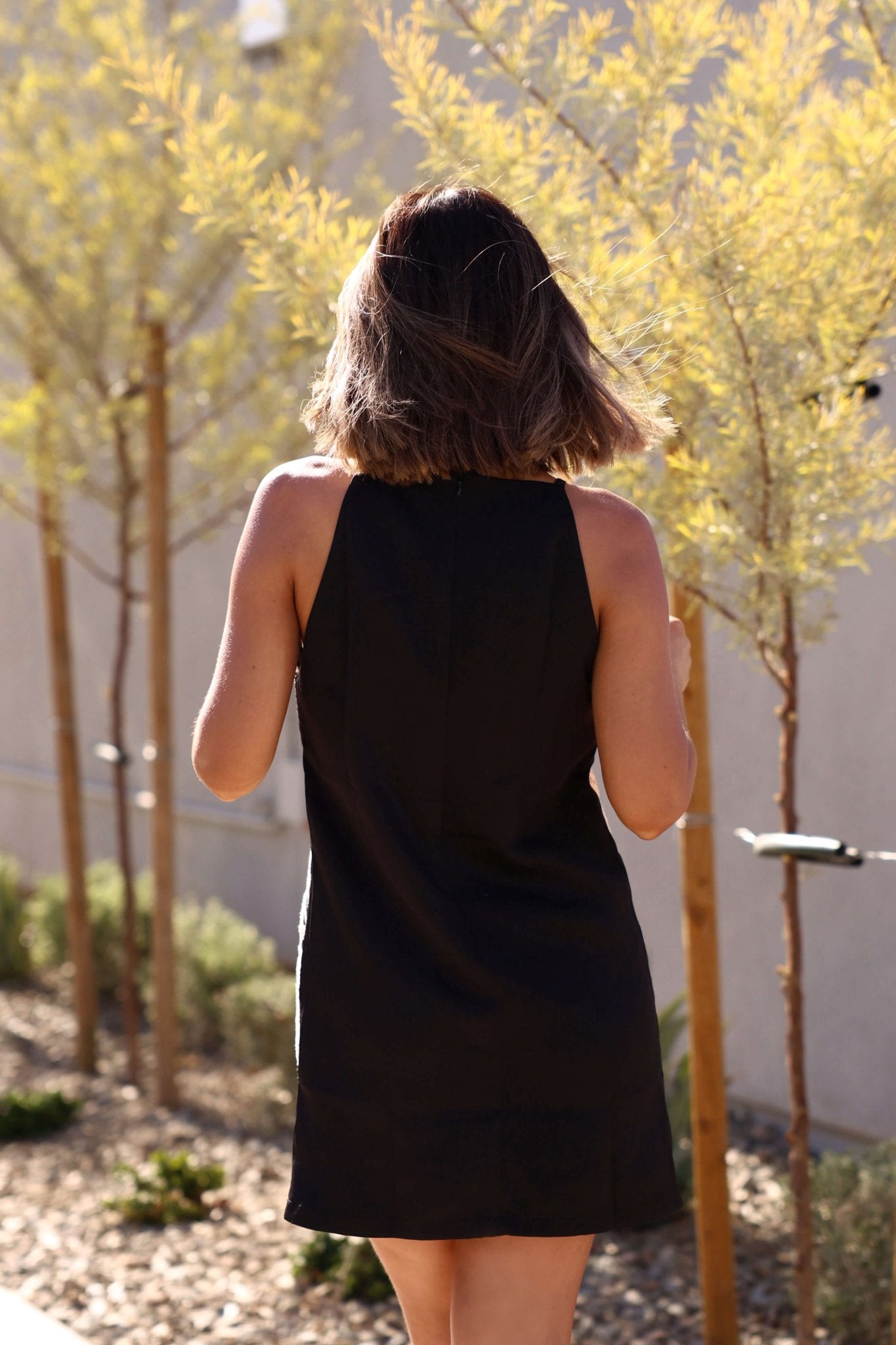 A woman in the Love & Lace Black Shift Mini Dress stands outdoors with her back to the camera on a sunny day.