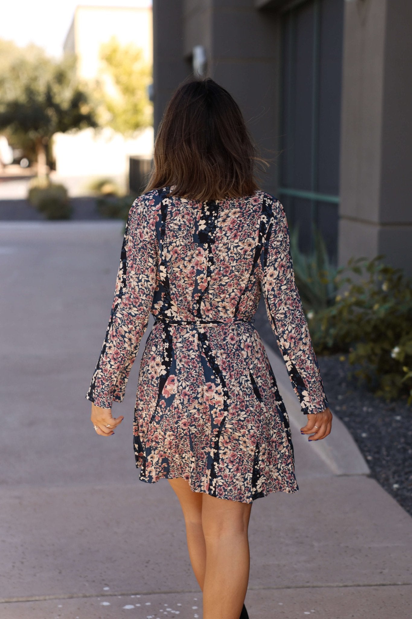 Woman with brown hair walks away in the Love Stitch Navy Floral Print Belted Mini Dress - FINAL SALE by a modern building.