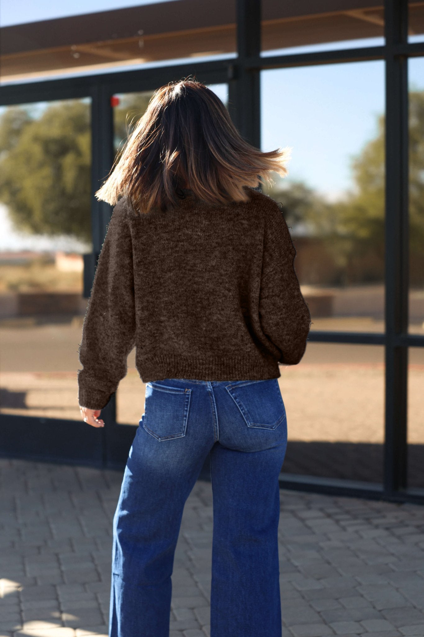 A person with shoulder-length hair, in blue jeans and the Lucille Brown Button Down Sweater Cardigan, stands outside facing away.