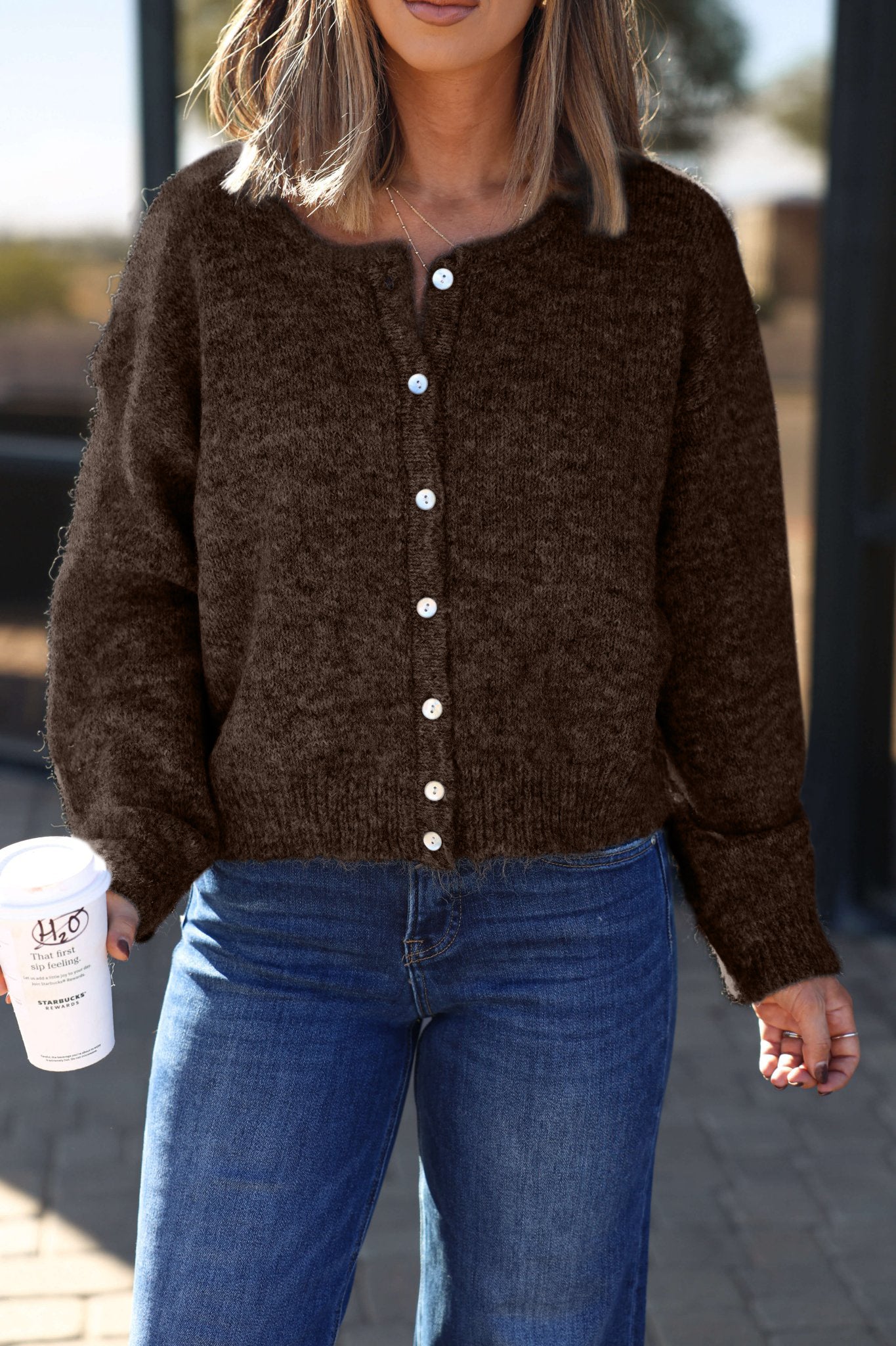 A woman wears the Lucille Brown Button Down Sweater Cardigan and blue jeans, holding a coffee cup outdoors, her face partially hidden.
