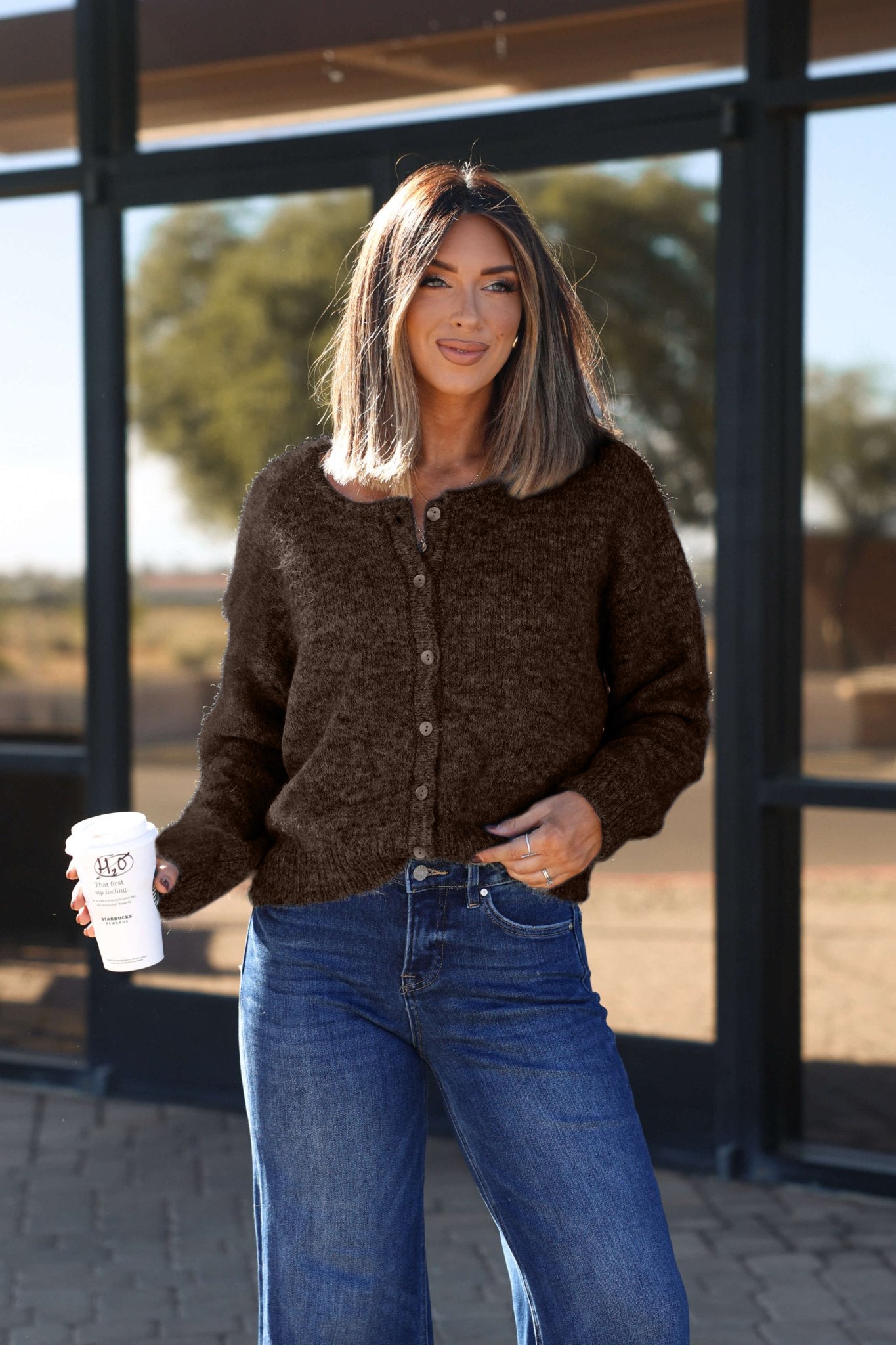 Woman with shoulder-length hair stands outside smiling, wearing the Lucille Brown Button Down Sweater Cardigan and jeans, holding coffee.