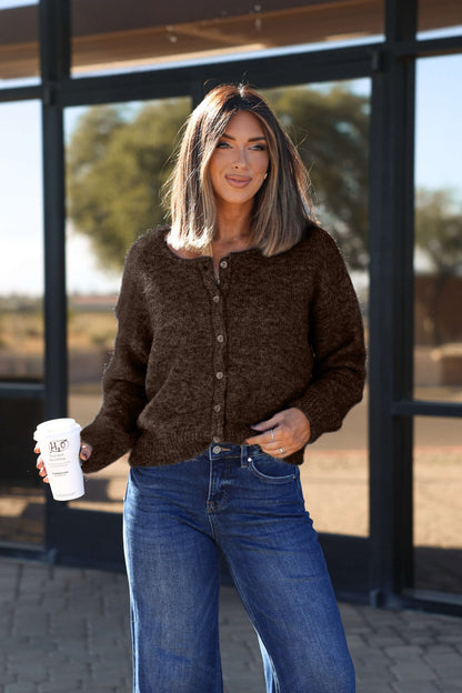 Woman with shoulder-length hair stands outside smiling, wearing the Lucille Brown Button Down Sweater Cardigan and jeans, holding coffee.