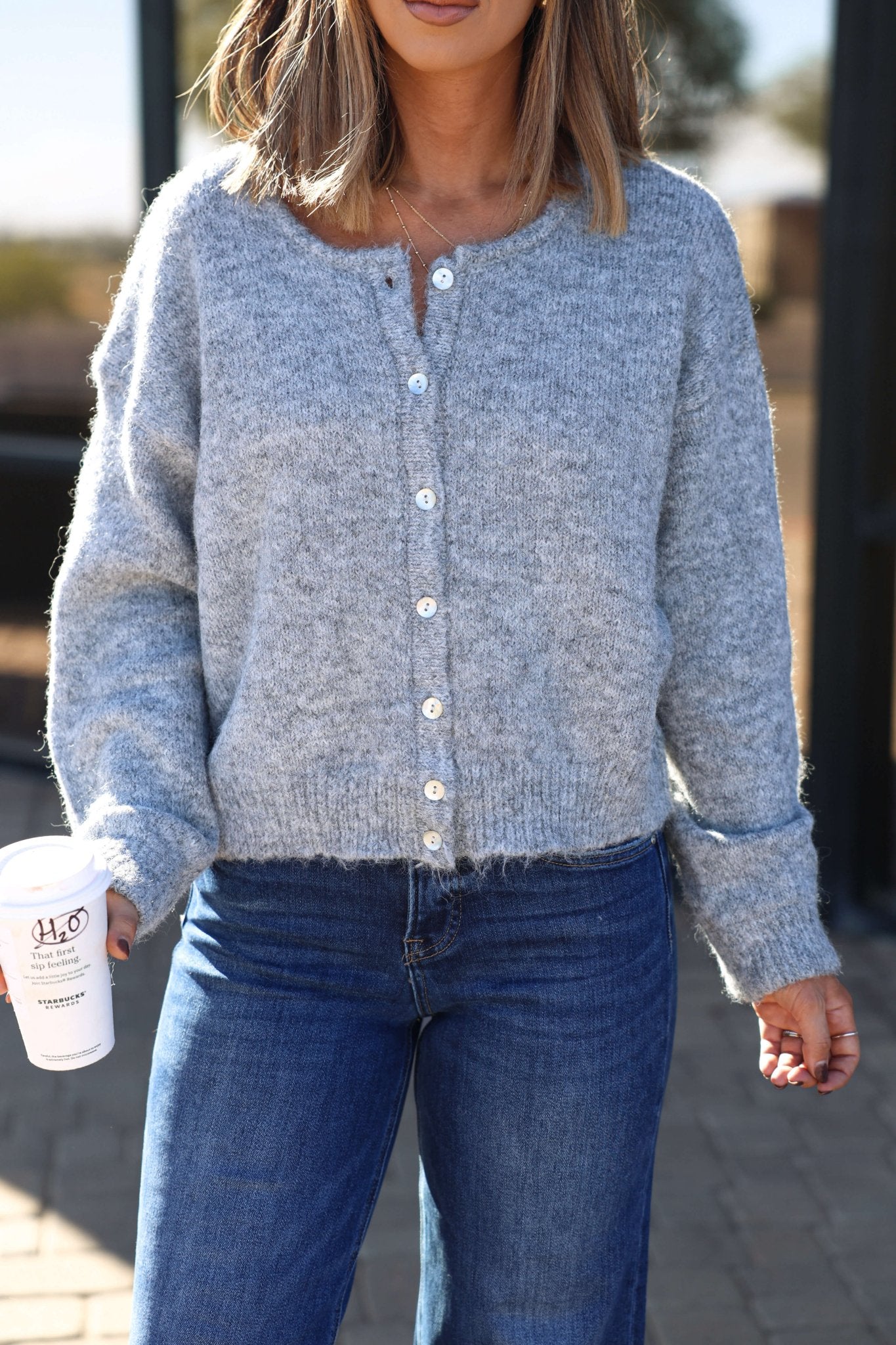 Woman wearing the Lucille Grey Button Down Sweater Cardigan and blue jeans, holding a white coffee cup outdoors.