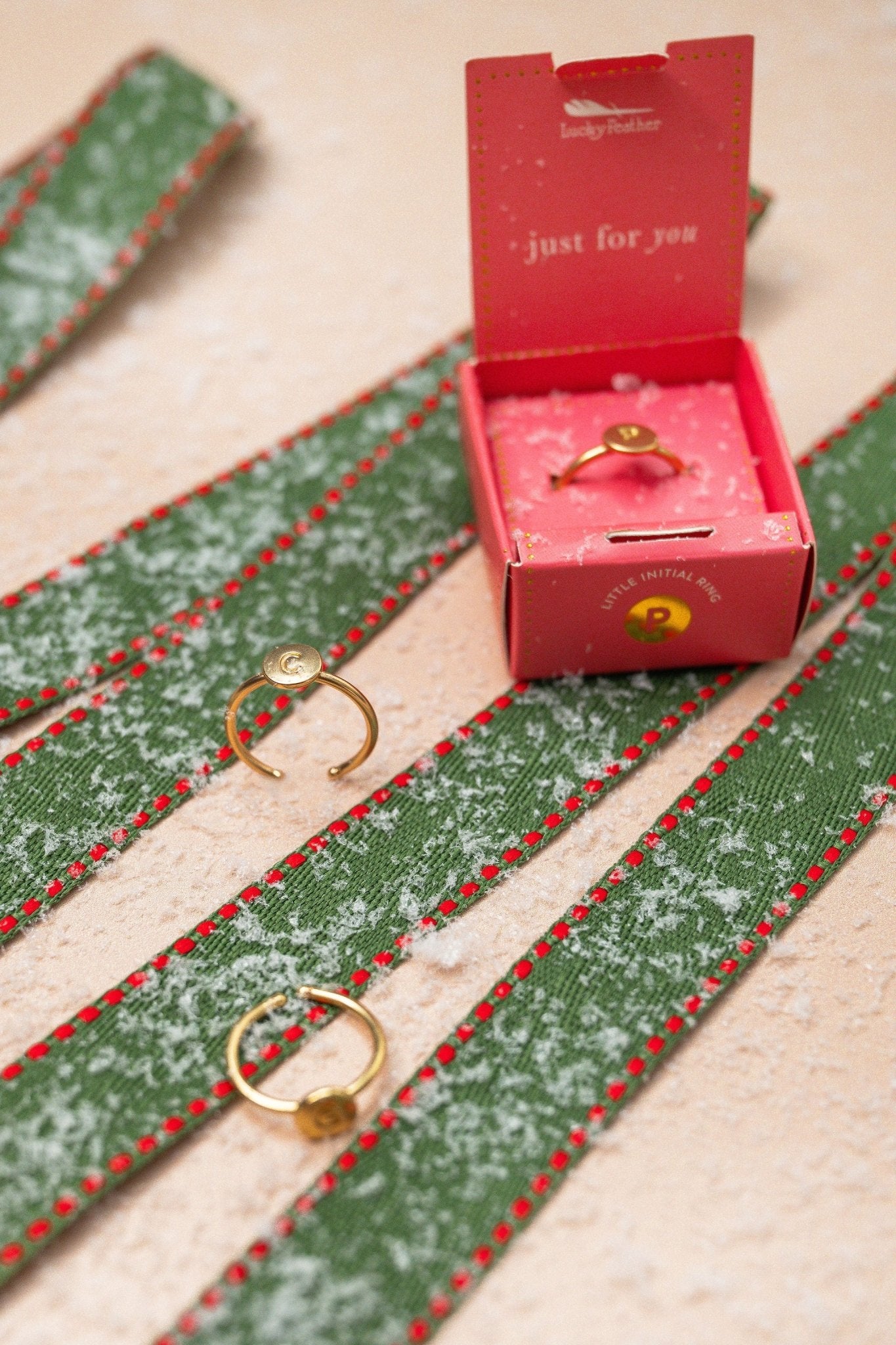 Lucky Feather Gold Initial Pendant Ring - FINAL SALE and pink jewelry box on green, snow-dusted ribbon with red stitching.