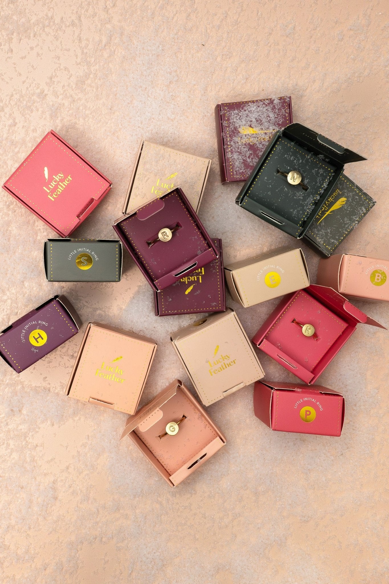 Lucky Feather Gold Initial Pendant Ring on pink textured surface with colorful jewelry boxes and gold initial pins.