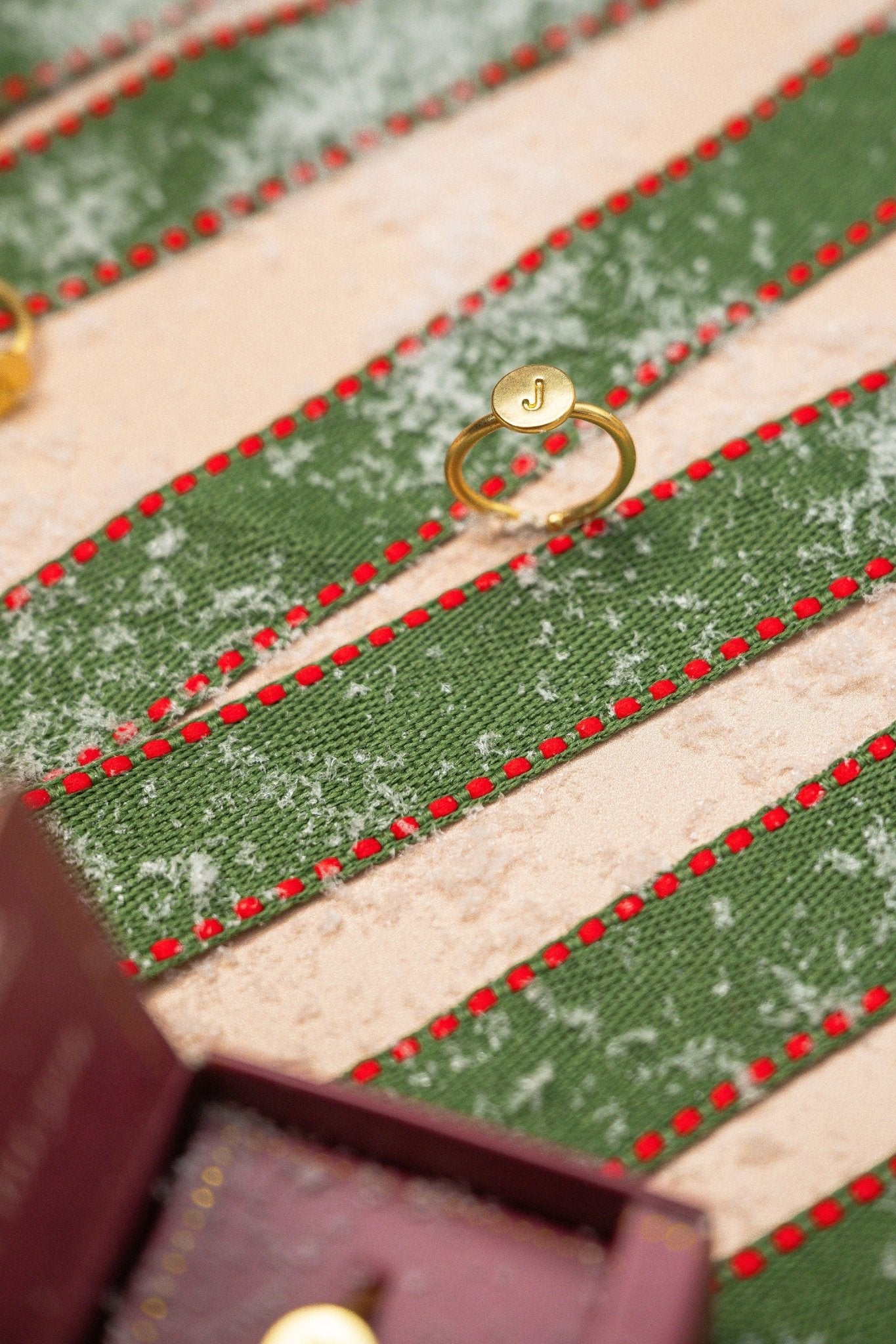 The Lucky Feather Gold Initial Pendant Ring - FINAL SALE with a J engraving rests on striped green and red snowy fabric.
