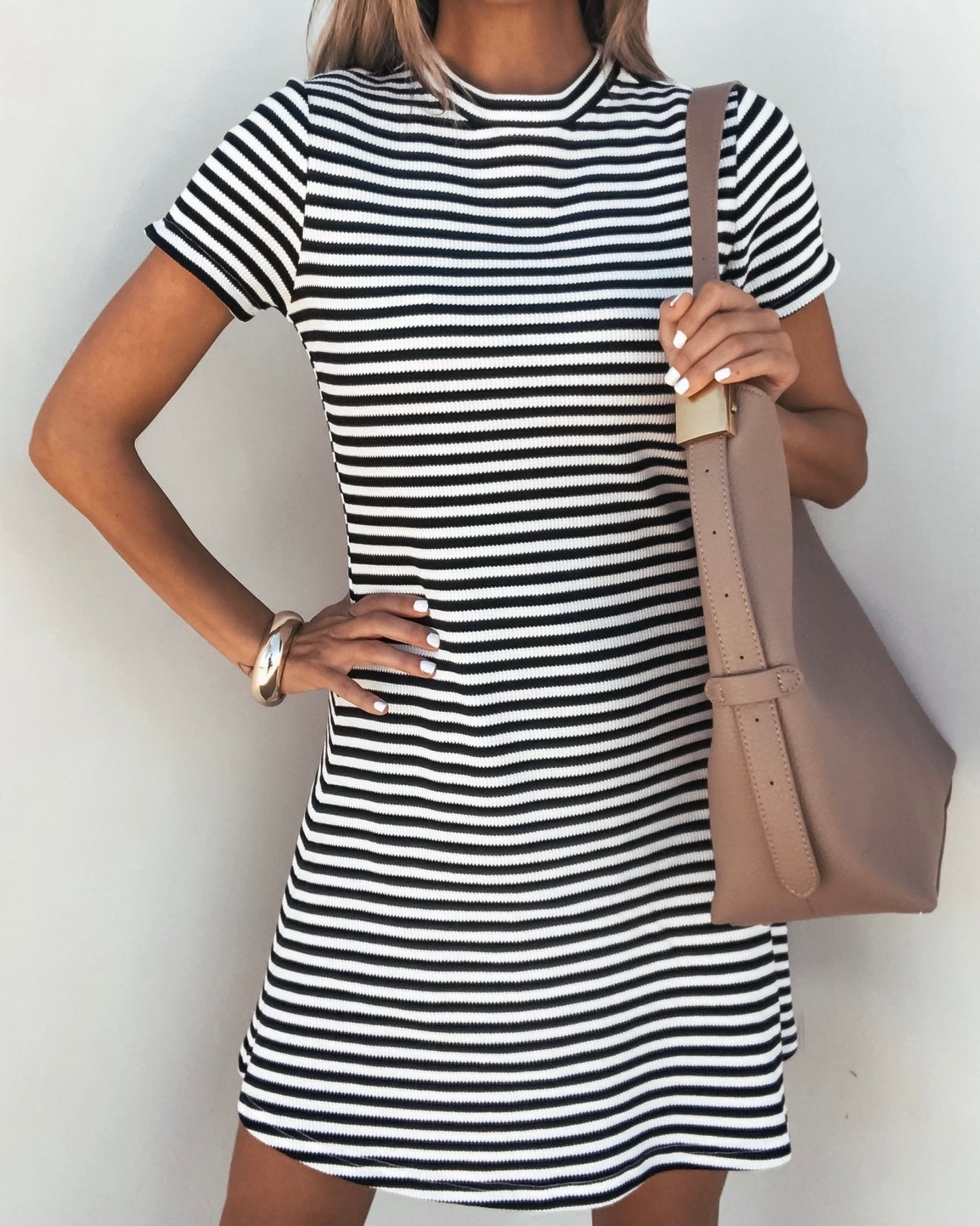 Model wears the Lucy Black and White Striped Mini Dress, holding a tan shoulder bag with hand on hip against a light background.