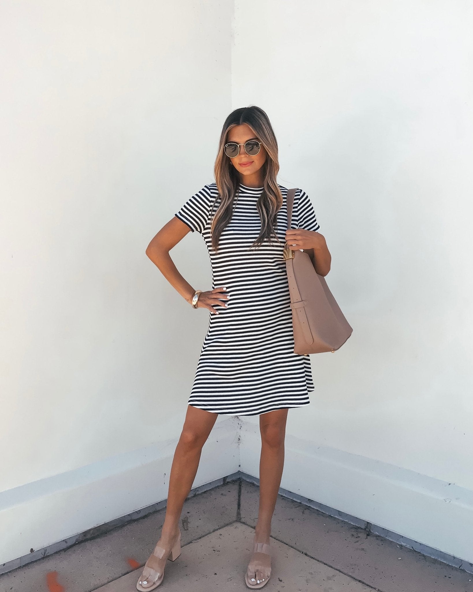 Woman wearing the Lucy Black and White Striped Mini Dress stands against a white wall, accessorized with sunglasses and a large handbag.