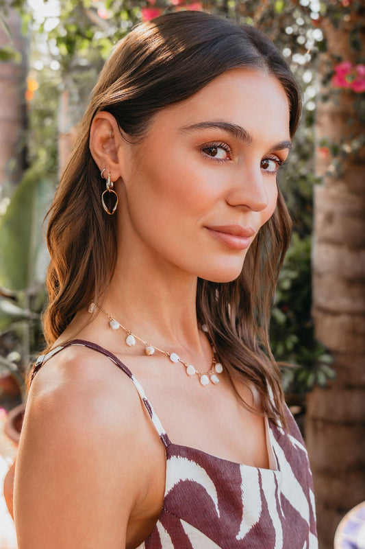A woman with long brown hair wears Luna Double Hoop Earrings, posing outdoors among green plants and pink flowers.