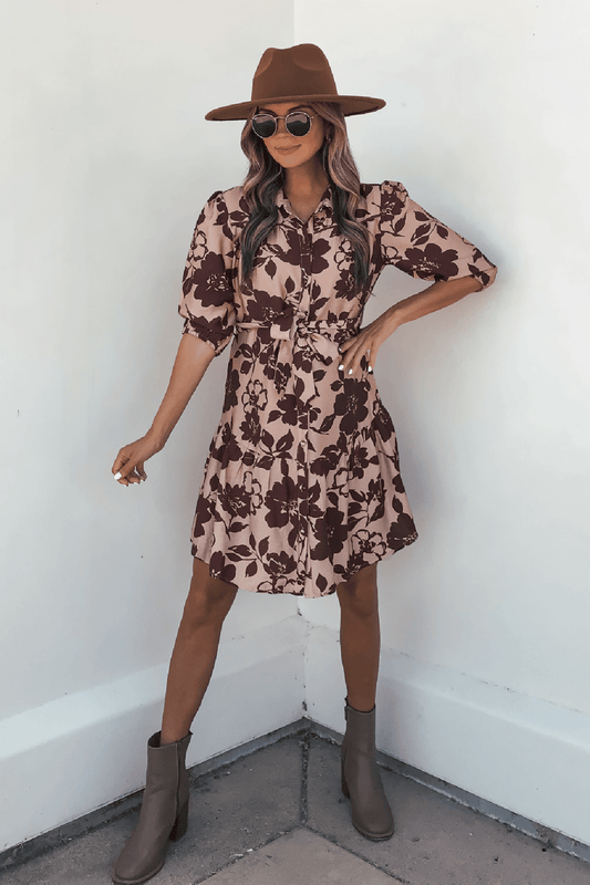Woman wearing the Lydia Brown Print Tie Front Mini Dress - DOORBUSTER poses with one hand on her hip against a white wall.