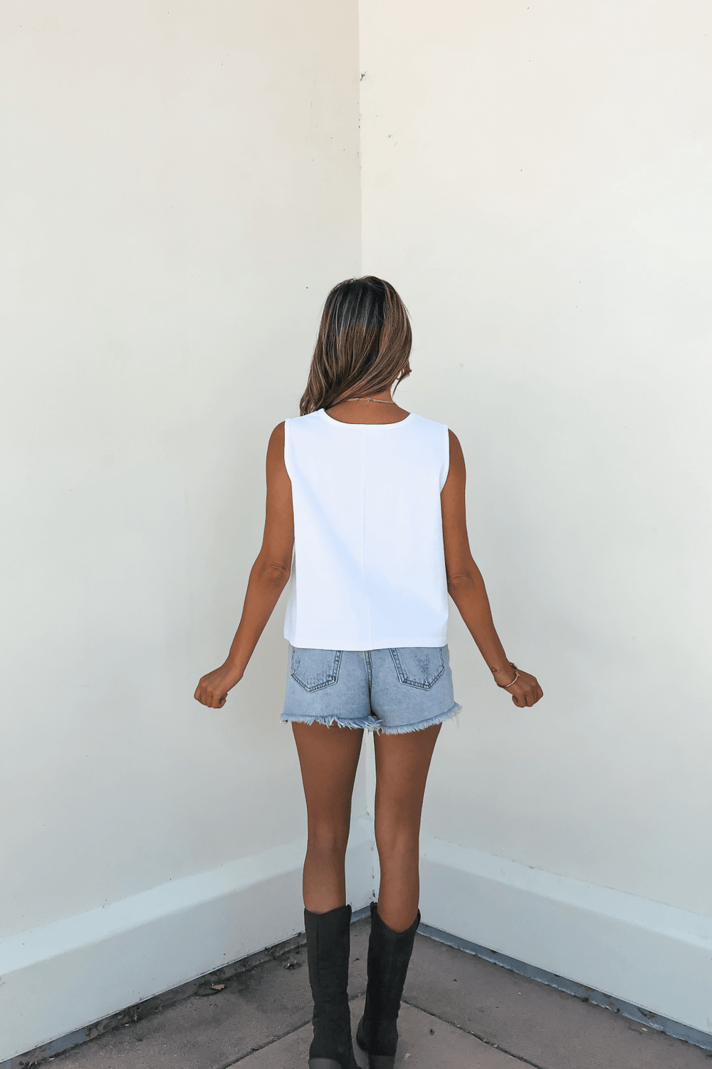 A woman stands in a corner wearing the Lydia White Crew Neck Tee, denim shorts, and black boots.