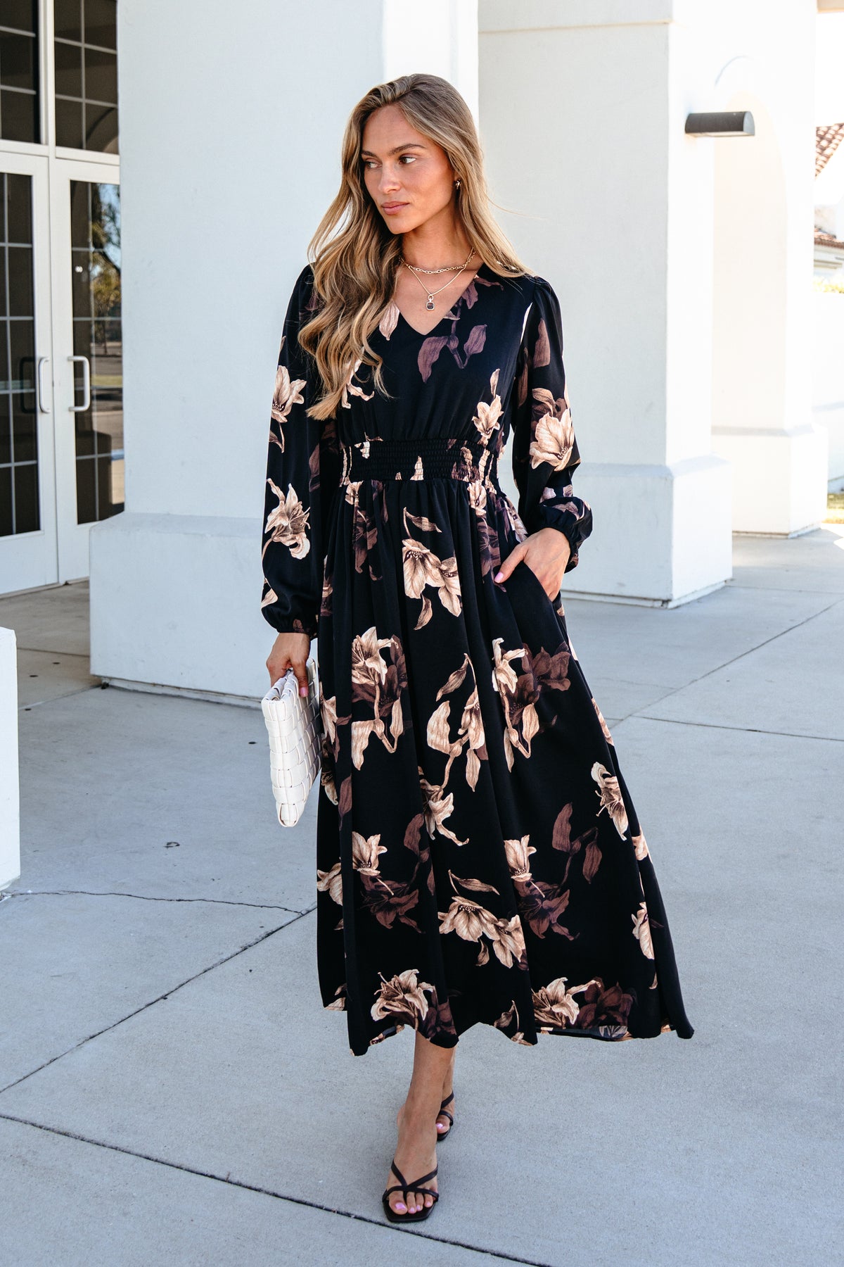 A woman wears a black Falling Leaves Print Maxi Dress with a tan floral print and a smocked waist, holding a white clutch.