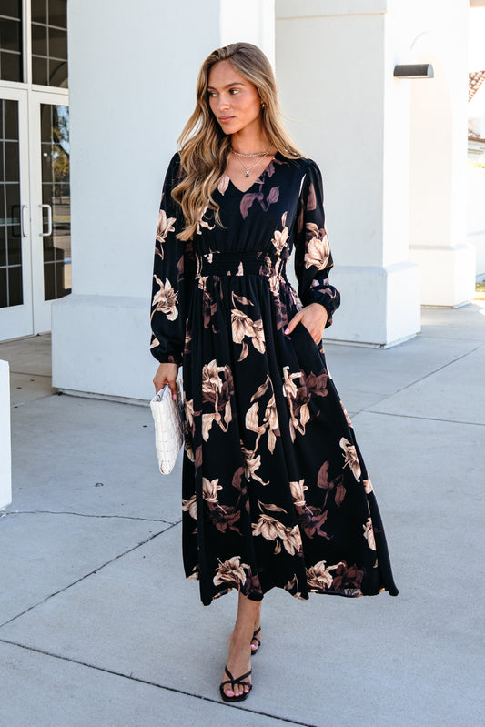 A woman wears a black Falling Leaves Print Maxi Dress with a tan floral print and a smocked waist, holding a white clutch.