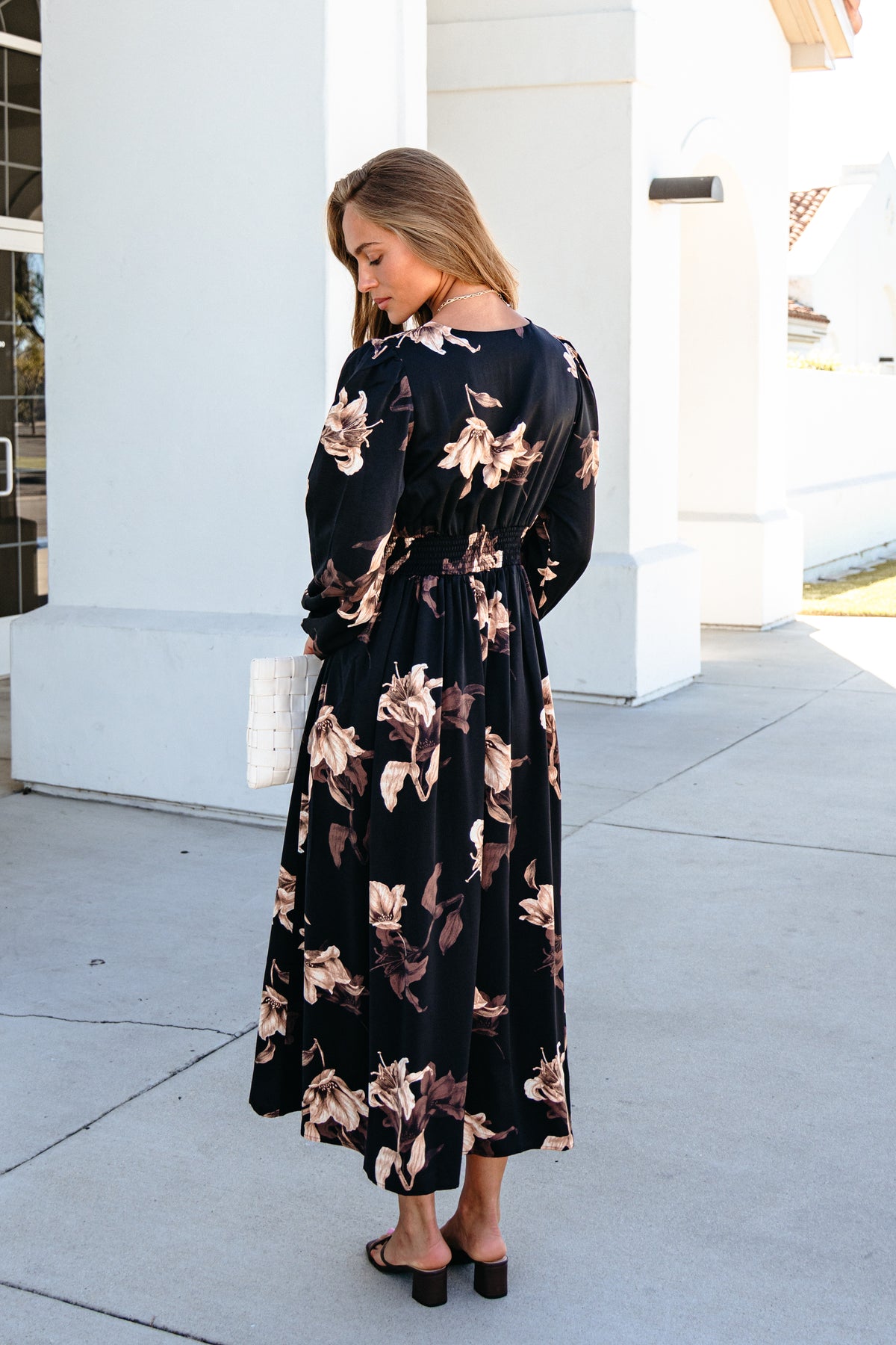 A person wearing a Falling Leaves Print Maxi Dress - Black with a smocked waist and long sleeves, holding a white woven handbag.