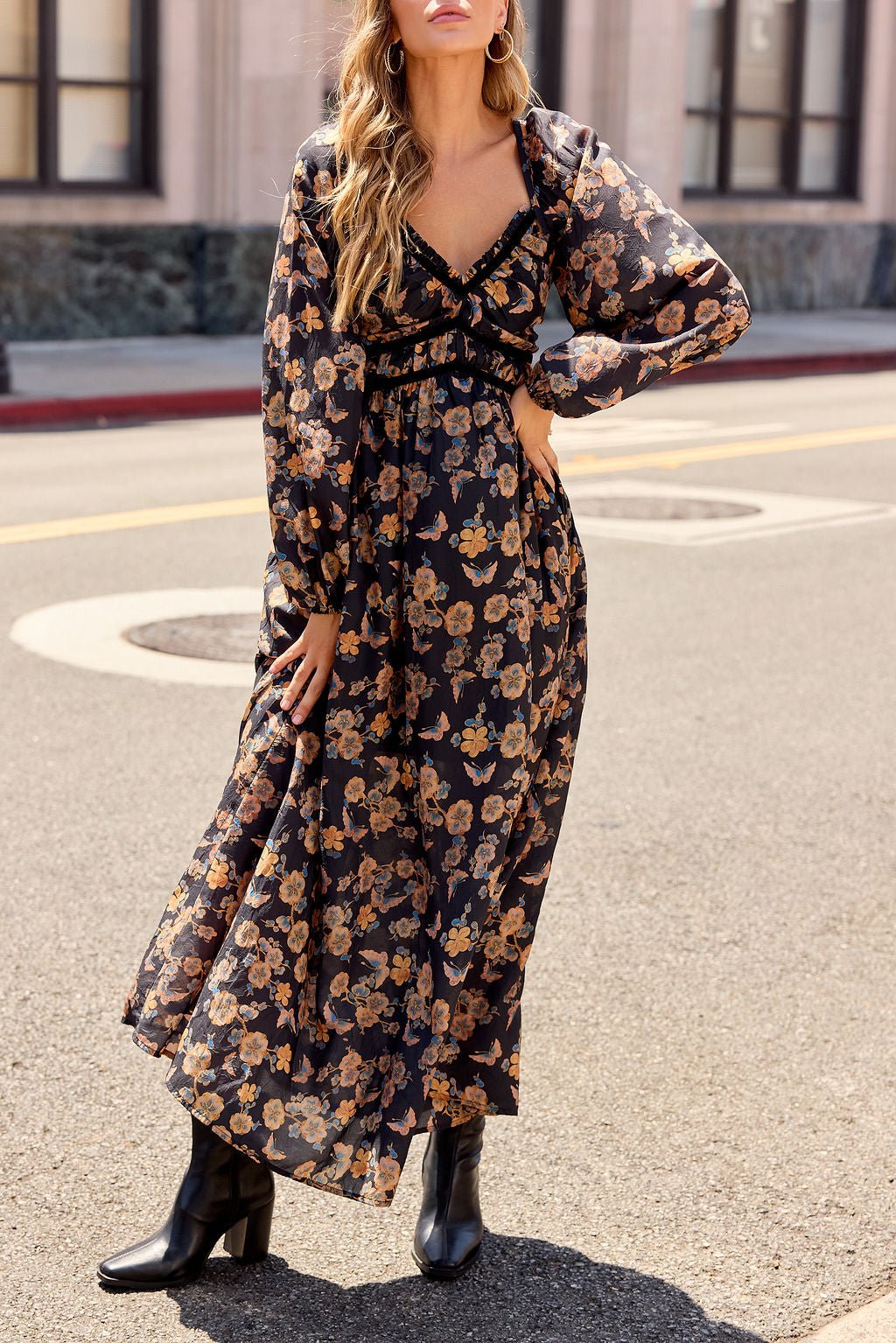 Woman in the Maple Floral Maxi Dress - Black and boots stands on a city street, hand on her hip.