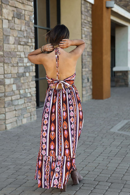A woman in the Maple Moments Halter Maxi Dress - Multi - DOORBUSTER walks on a stone path, adjusting her hair with both hands.