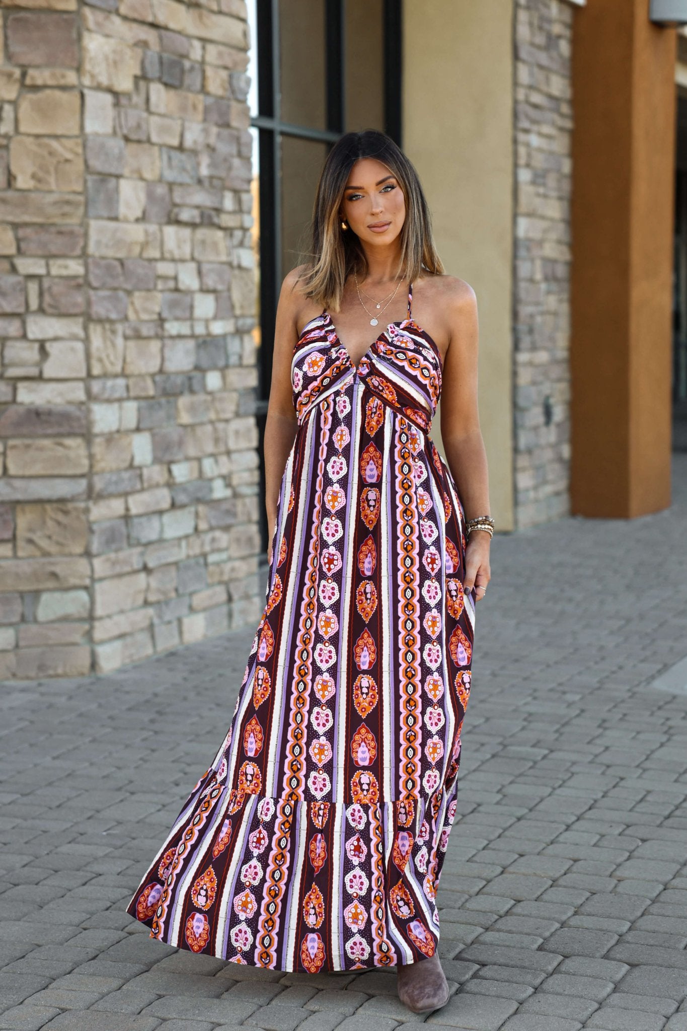 Woman in the Maple Moments Halter Maxi Dress - Multi stands on a paved walkway near a stone and stucco building.