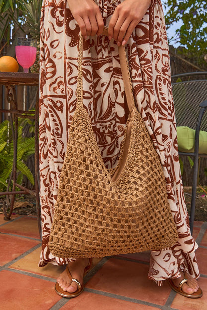 Wearing a patterned brown and white dress, she holds a Marbella Khaki Tote while standing on a tiled patio near plants and a drink.