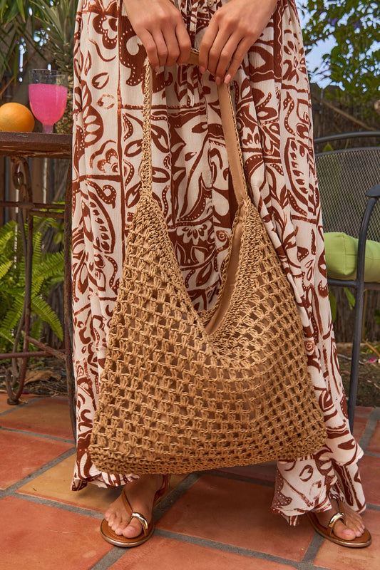 Wearing a patterned brown and white dress, she holds a Marbella Khaki Tote while standing on a tiled patio near plants and a drink.