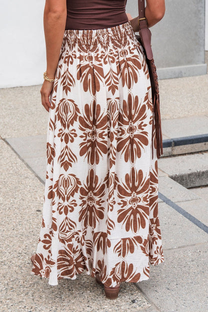 Someone in the Marchesa Print Maxi Skirt - Taupe stands on a pebble sidewalk.
