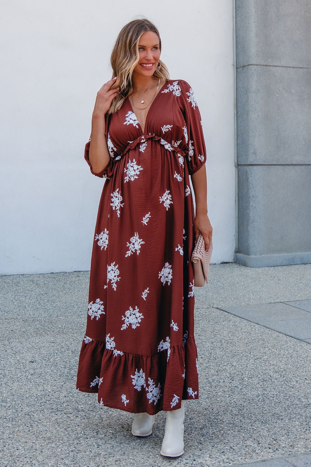 Woman in the Maribel Brown Floral Maxi Dress - FINAL SALE, white boots, and a beige clutch stands outside by a light wall.