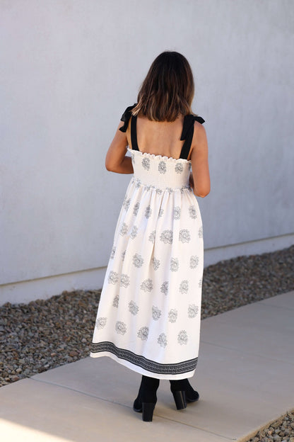 Woman stands outdoors in the Maribel Cream Border Print Smocked Midi Dress - DOORBUSTER by a stone border.