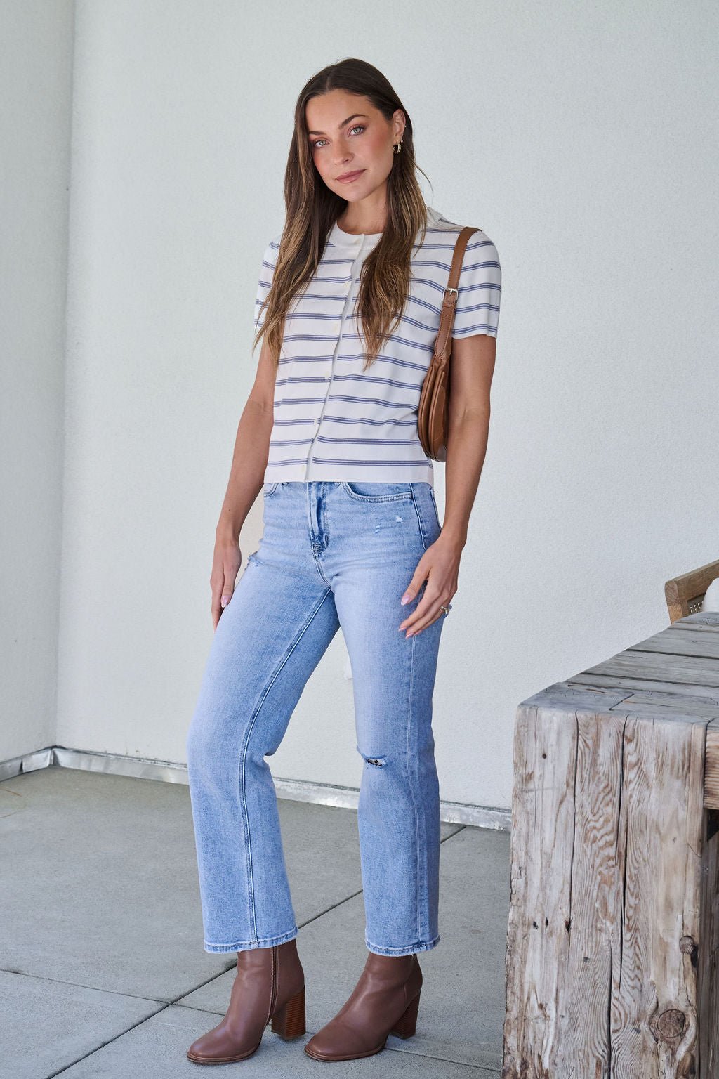 A woman in a Marina Blue and White Striped Sweater Cardigan, light blue jeans, and brown boots holds a brown purse by a wooden table.