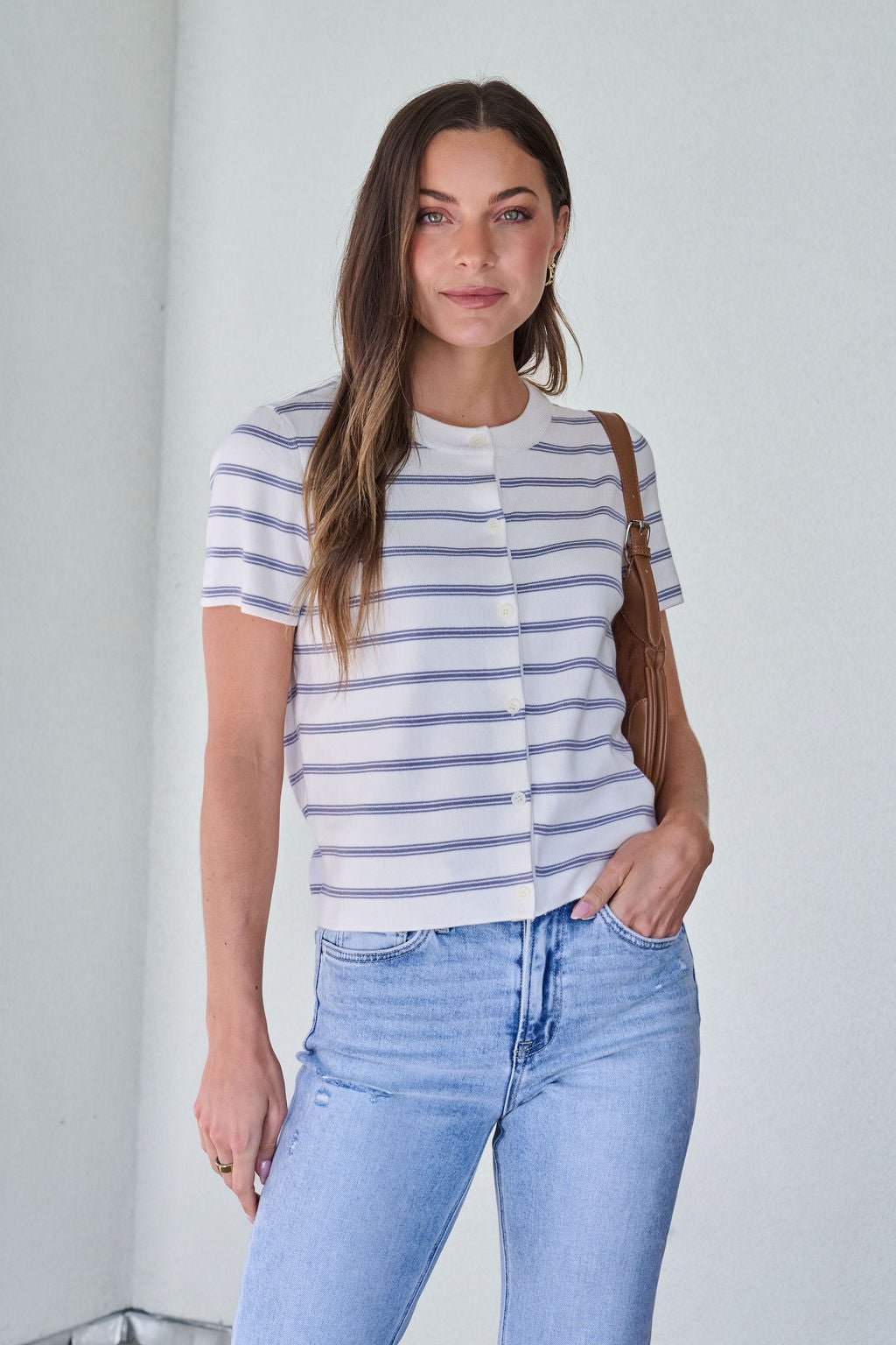 Woman in the Marina Blue and White Striped Sweater Cardigan, light blue jeans, and a brown bag stands indoors.
