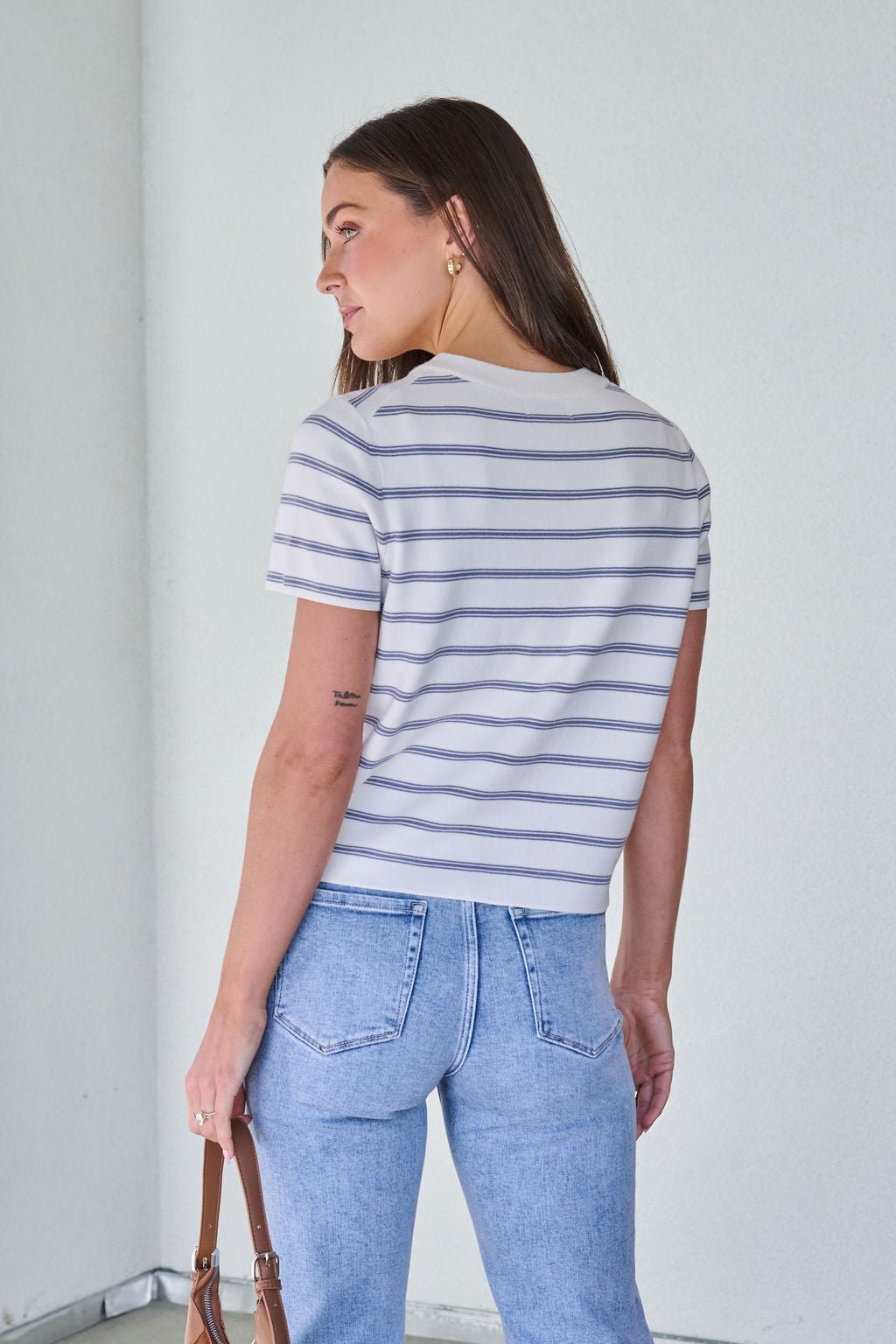 Woman with long brown hair in a Marina Blue and White Striped Sweater Cardigan and light blue jeans, indoors holding a brown bag.