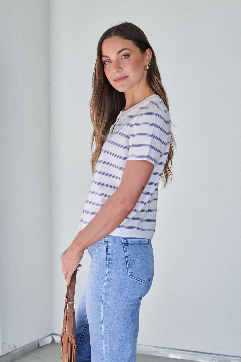 Woman with long brown hair wears a Marina Blue and White Striped Sweater Cardigan, light blue jeans, and holds a tan bag, smiling.