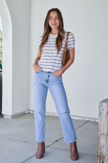 Woman stands smiling in a Marina Blue and White Striped Sweater Cardigan, ripped light-blue jeans, brown boots, and a brown shoulder bag.