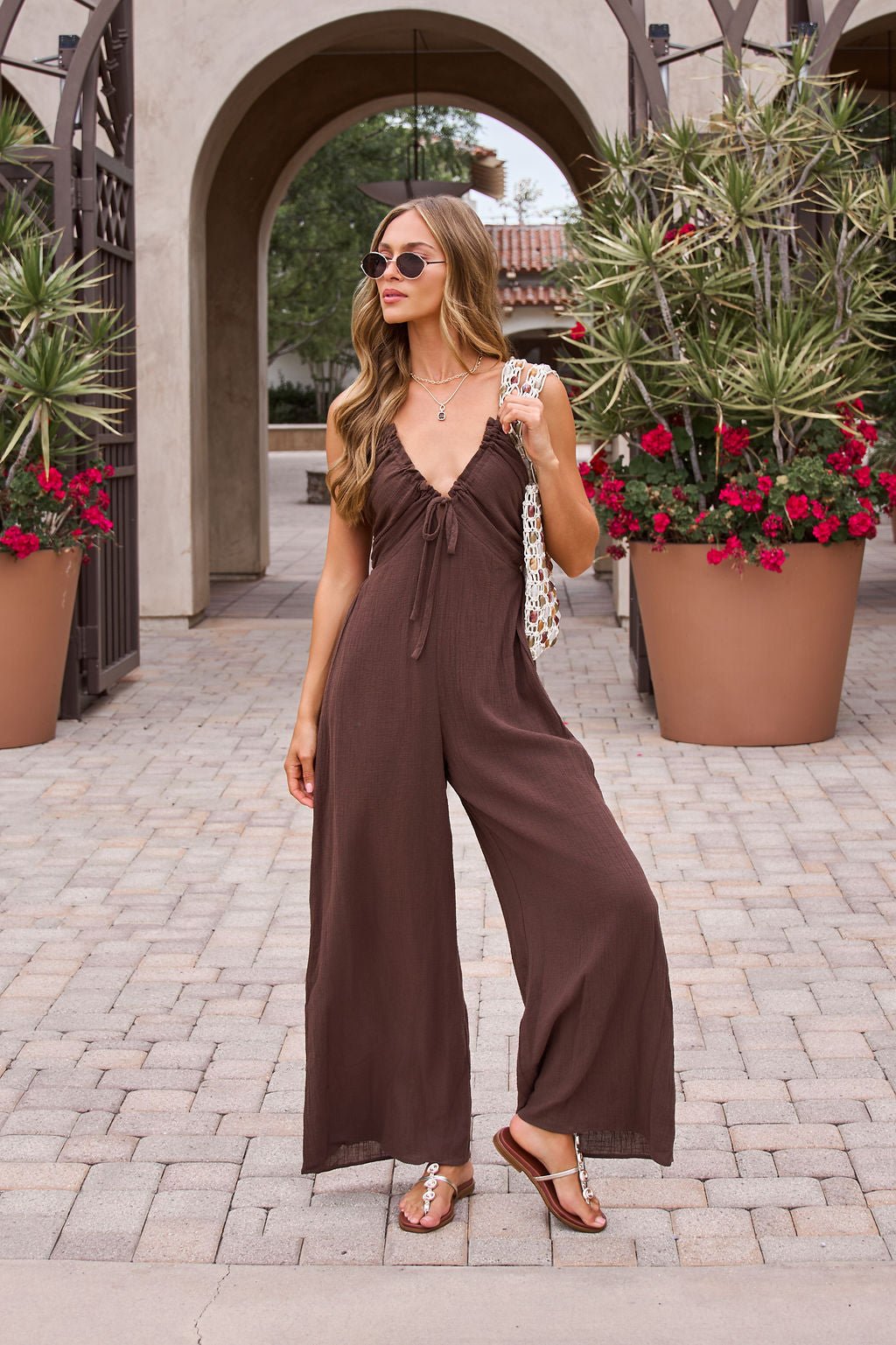 Woman in a Marisol Brown Tie‑Front Wide Leg Jumpsuit, sandals, and sunglasses stands near potted plants with red flowers.