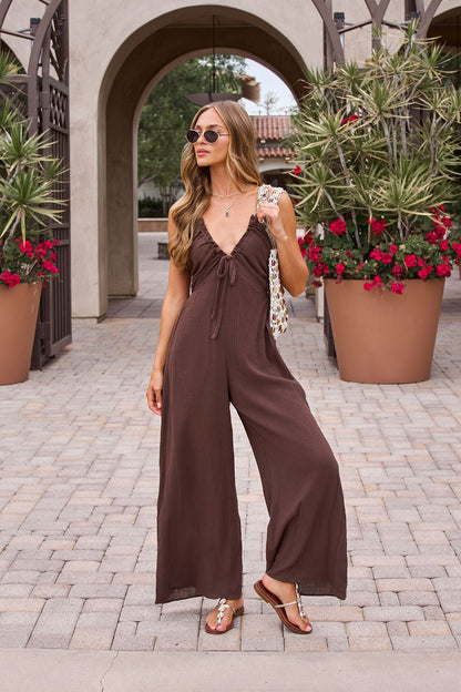 Woman in a Marisol Brown Tie‑Front Wide Leg Jumpsuit, sandals, and sunglasses stands near potted plants with red flowers.