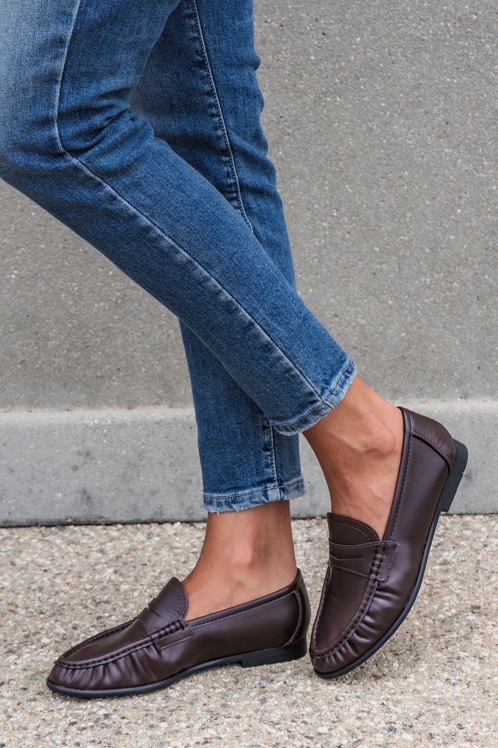 Individual in blue jeans and brown Matisse Carlton Loafers stands on concrete with legs crossed.