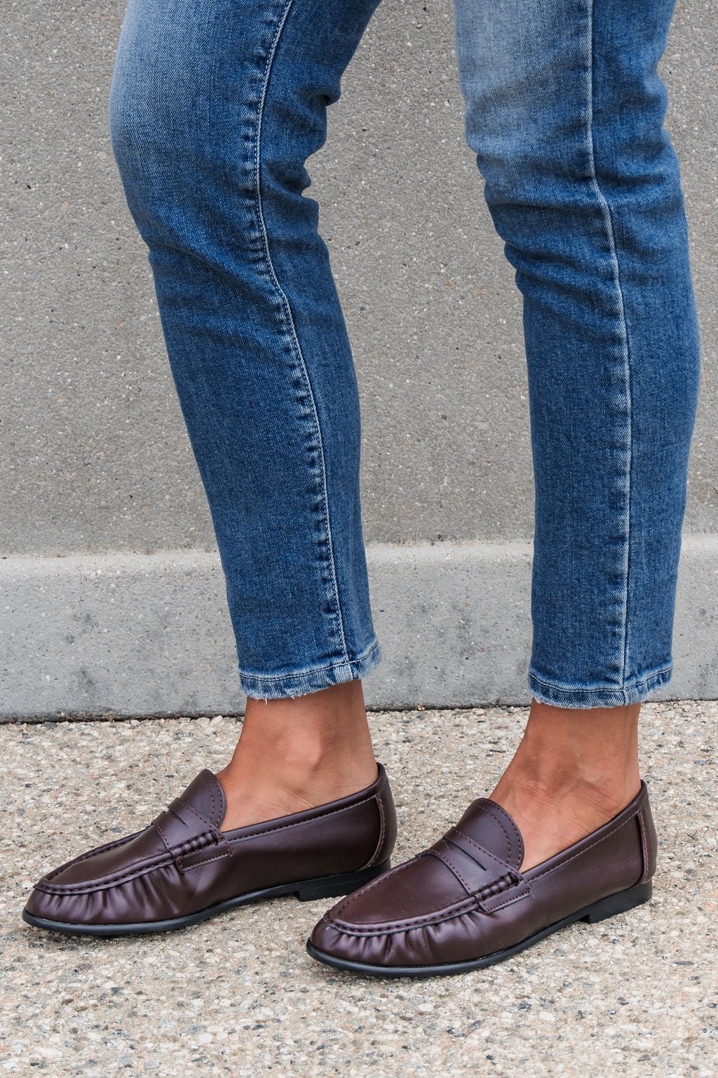 Person in blue jeans and Matisse Carlton Loafers - Brown stands on gravel before a concrete wall.