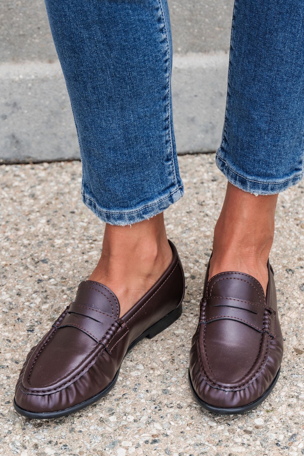 Individual in Matisse Carlton Loafers - Brown and cropped blue jeans stands on a gravel sidewalk.