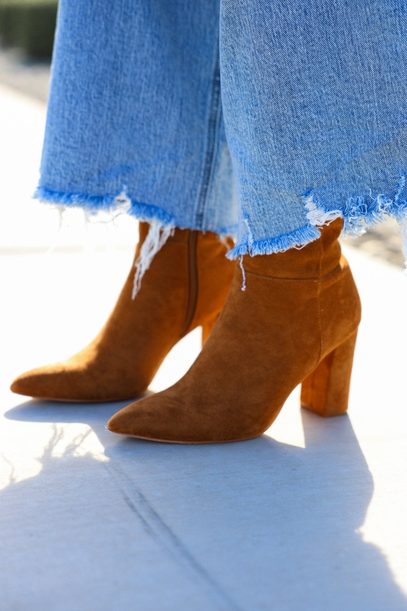 A person in frayed hem blue jeans and Matisse Spencer Camel Heeled Ankle Booties stands on a sunny sidewalk.