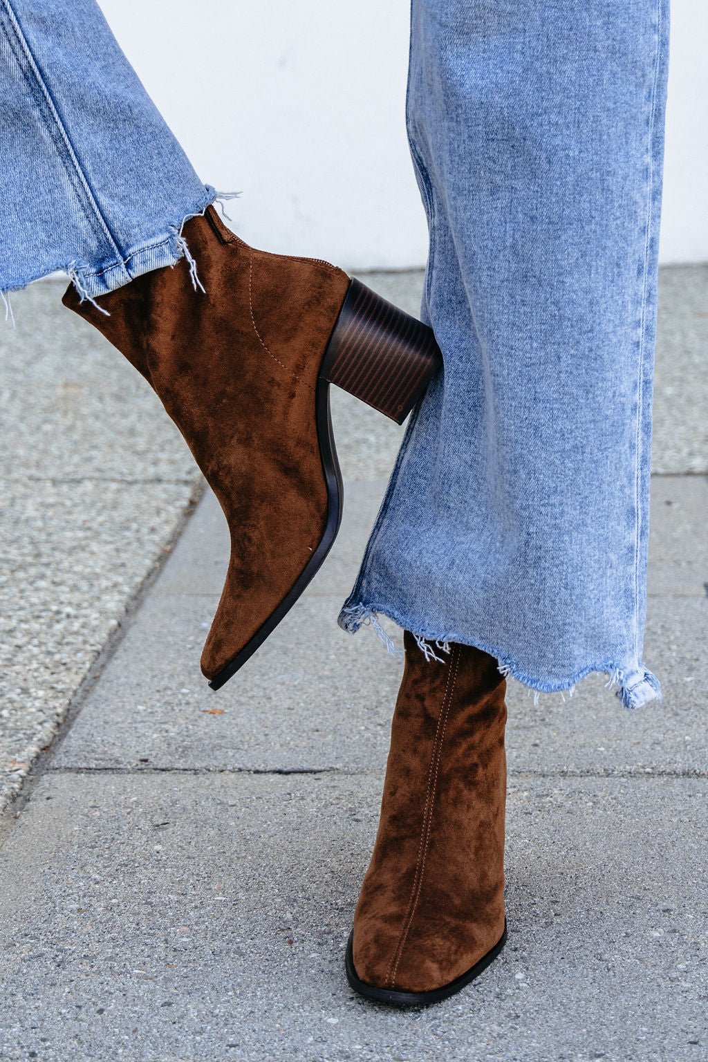Person in Matisse Warren Heeled Ankle Booties - Brown and frayed hem jeans stands on a sidewalk, one foot crossed over the other.