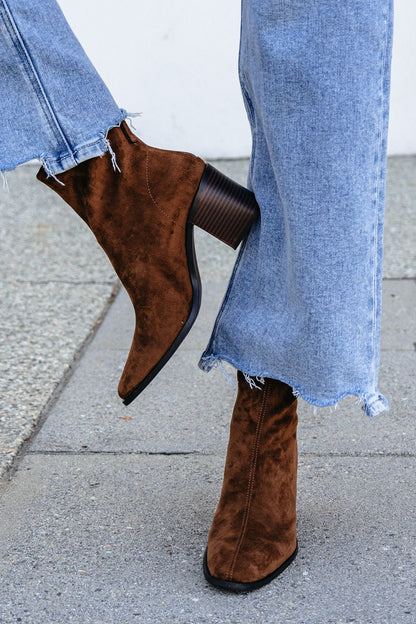 Person in Matisse Warren Heeled Ankle Booties - Brown and frayed hem jeans stands on a sidewalk, one foot crossed over the other.