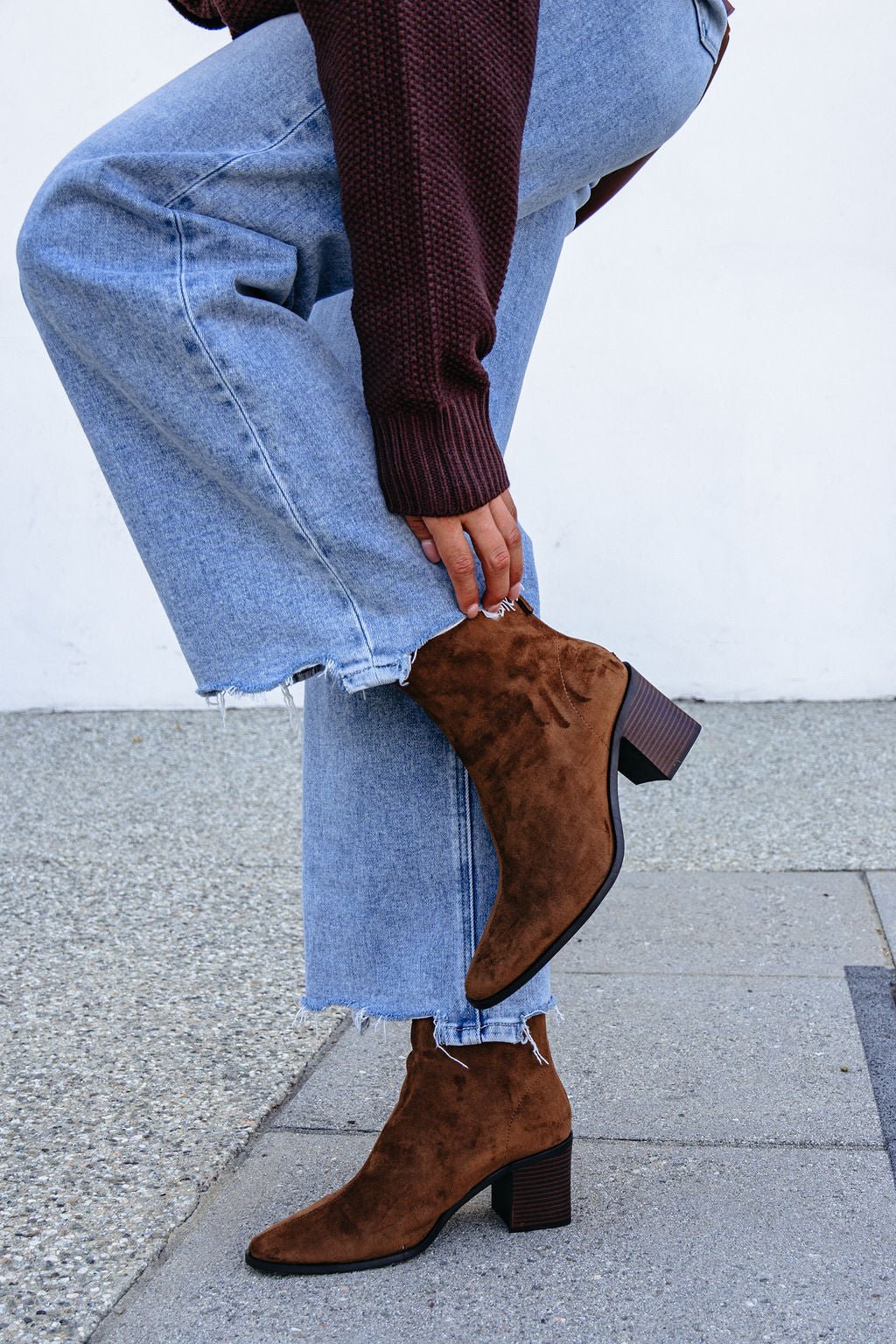 Person in Matisse Warren Heeled Ankle Booties - Brown and frayed light-wash jeans bends down stylishly on the sidewalk.