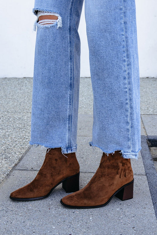 Person in light blue ripped jeans and Matisse Warren Heeled Ankle Booties - Brown, standing stylishly on a stone sidewalk.