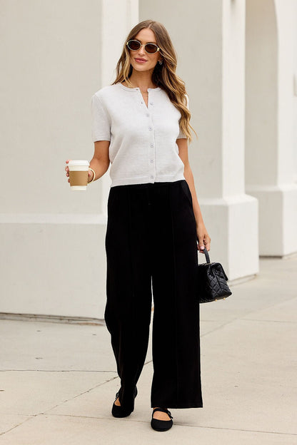 Woman wearing sunglasses, the Maura Grey Short Sleeve Sweater Cardigan, black wide-leg pants, holding coffee and a small bag outside.