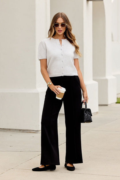 Woman in sunglasses, Maura Grey Short Sleeve Sweater Cardigan, black pants, and flats holds a coffee cup and handbag outside.