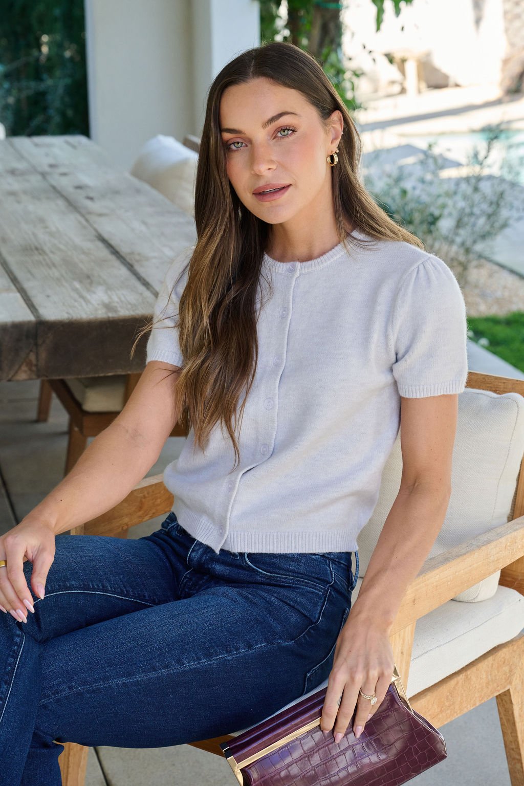 Woman with long brown hair in a Maura Grey Short Sleeve Sweater Cardigan and jeans, sitting on a patio chair holding a purple clutch.