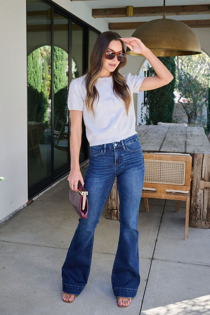 Woman wearing sunglasses, Maura Grey Short Sleeve Sweater Cardigan, flared jeans, and a clutch stands by a rustic wooden table outside.
