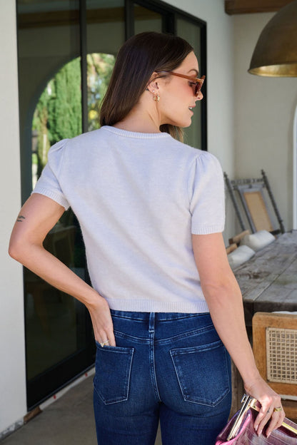 A woman in sunglasses wears the Maura Grey Short Sleeve Sweater Cardigan outdoors, facing away and holding a metallic purse.