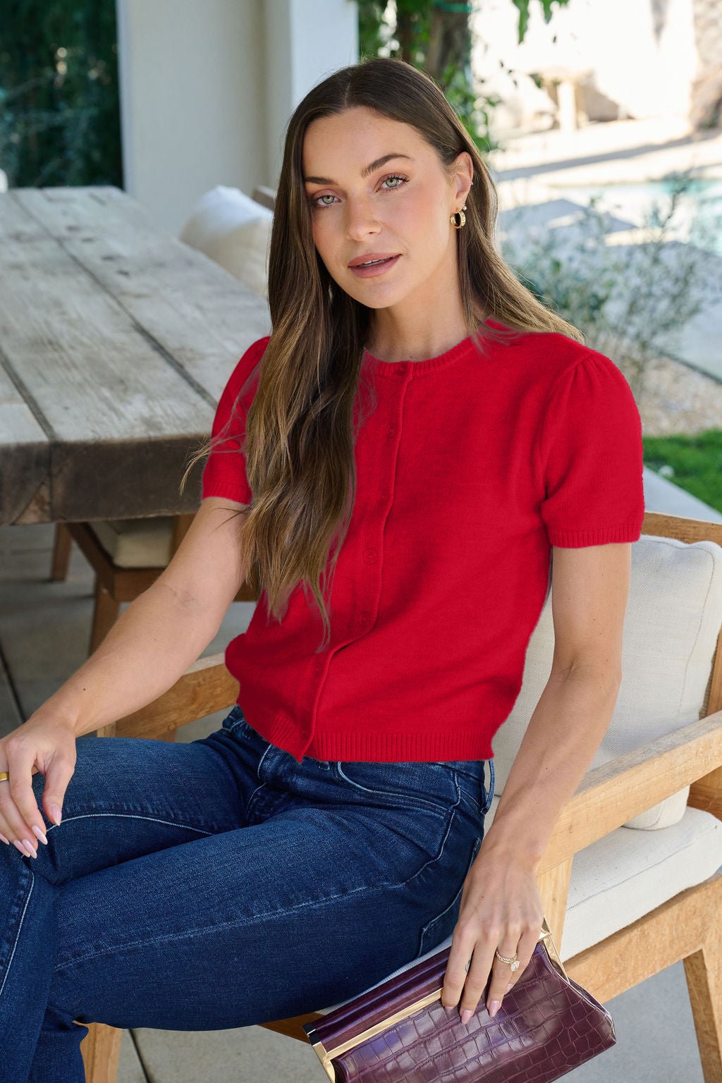 Woman in Maura Red Short Sleeve Sweater Cardigan sits on a chair outdoors, holding a brown clutch by a wooden table.